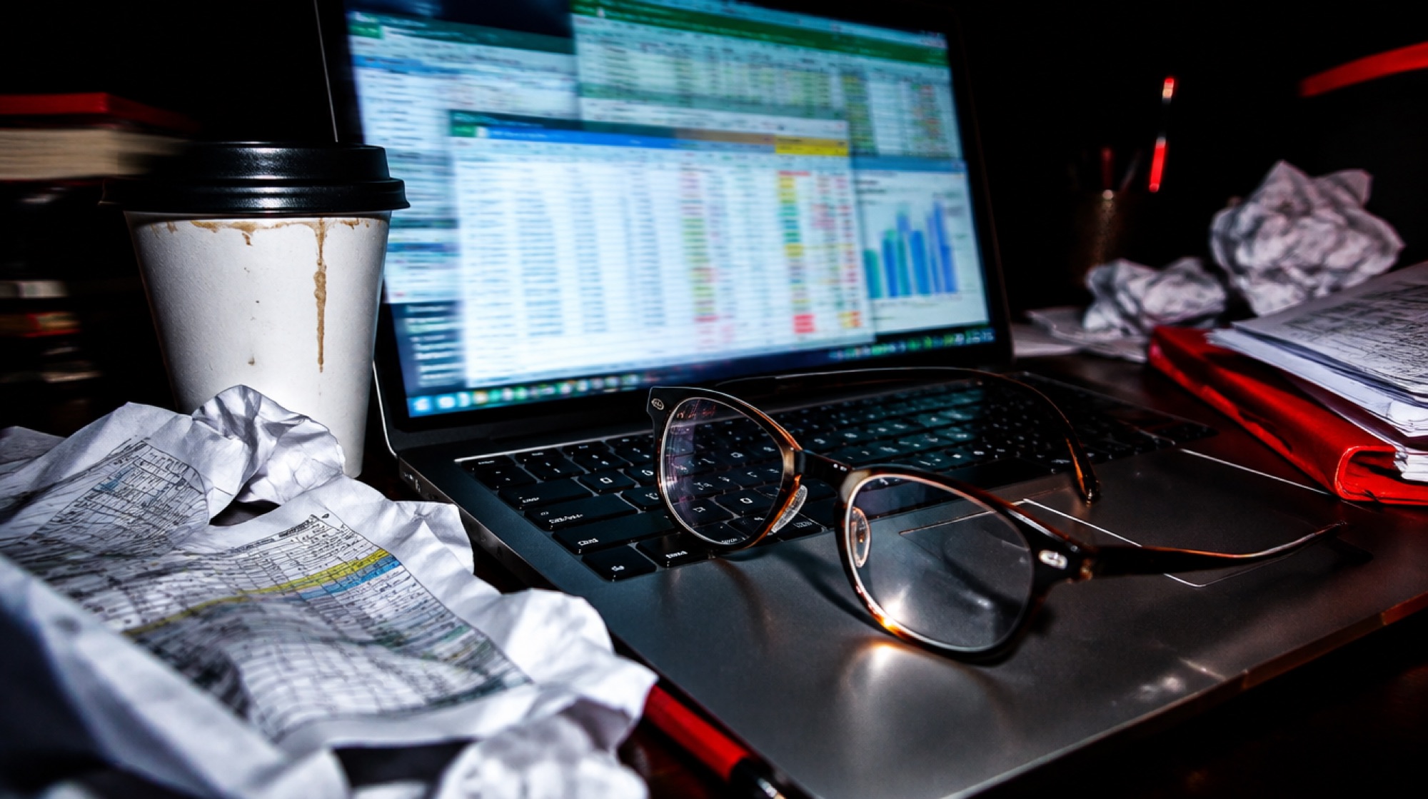 Glasses resting on a laptop keyboard at night, the screen full of spreadsheet windows, a cold coffee beside it.